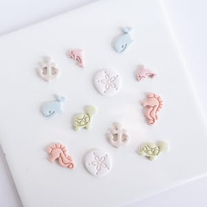 Könnte beinhalten: Eine Sammlung von pastellfarbenen Verzierungen zum Thema Meer, darunter Anker, Delfine, Wale, Seepferdchen, Schildkröten und Sanddollar. Die dekorativen Elemente sind auf einer weißen Oberfläche angeordnet und bieten ein maritimes Thema.