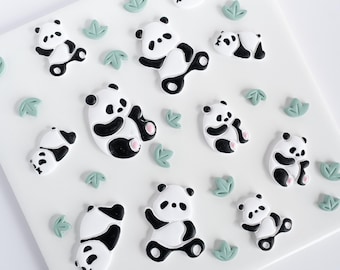 May include: A collection of small, flat, decorative panda bear figures in black and white with pink accents, arranged on a white surface alongside several tiny, light green, three-leaf plant shapes scattered throughout the composition.