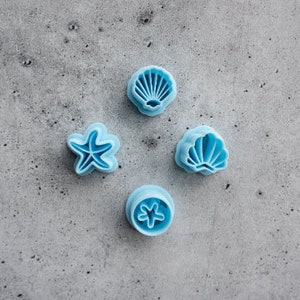 May include: Four light blue seashell and starfish shaped cookie cutters are arranged on a grey speckled surface. The cutters include two seashell designs, a starfish, and a circular cutter with a starfish imprint. The cutters are made of plastic.