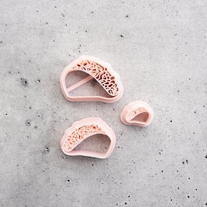 May include: Three light pink plastic cookie cutters in the shape of a taco. Two of the cutters are larger and feature a detailed, textured interior pattern resembling shredded lettuce or meat, while the third cutter is smaller and simpler.