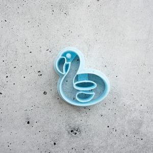May include: A light blue cookie cutter shaped like a flamingo. The tool features a defined outline of the bird's head, neck, and body, with an internal structure that creates the wing and beak details when pressed into dough or other soft materials.
