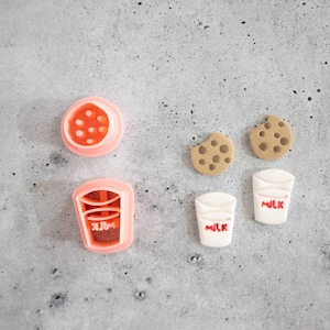 May include: A set of cookie cutters for making milk carton and chocolate chip cookie shapes. The cutters are pink and white plastic. The set includes two milk carton cutters and two chocolate chip cookie cutters.