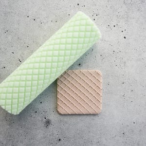 May include: A light green cylindrical roller with a diamond-shaped grid pattern and a square tan tile featuring a matching diamond-shaped grid pattern. Both items have a textured surface and are positioned diagonally on a grey speckled surface.