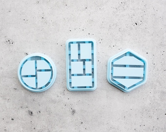 Puede incluir: Tres cortadores de galletas de plástico azul claro con formas circular, rectangular y hexagonal. Cada cortador presenta una rejilla interna o un diseño segmentado para crear patrones o impresiones específicas en la masa o arcilla.