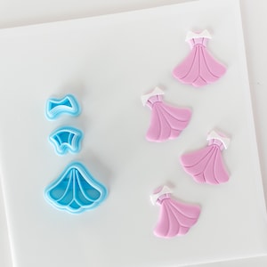 May include: A set of blue plastic cookie cutters shaped like a dress, a bow, and a collar, and four pink fondant dresses.