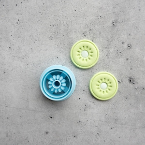 May include: A light blue circular object with a central flower-like pattern and two smaller, separate light green circular discs, each featuring a matching embossed flower design with a solid center and radiating petal-like indentations.