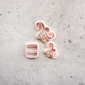 Pode incluir: Três cortadores de biscoitos de plástico rosa claro. Dois dos cortadores têm a forma de dinossauros com pescoços e caudas longos, enquanto o terceiro tem a forma de um copo cilíndrico com linhas horizontais no centro da forma.