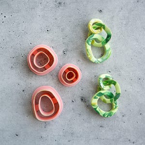 May include: Three pink plastic clay cutters with irregular oval shapes and two dangling earrings composed of interconnected lime green and dark green marbled rings. The cutters feature a double-walled design, while the earrings display a smooth finish.