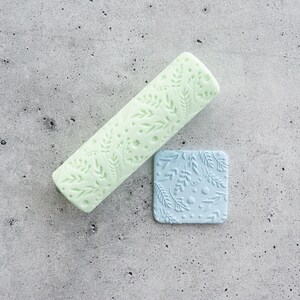 Pode incluir: Um rolo cilíndrico verde-claro com um padrão em relevo de folhas e pontos, acompanhado por um pequeno azulejo quadrado azul-claro que apresenta a mesma textura botânica em relevo na sua superfície.