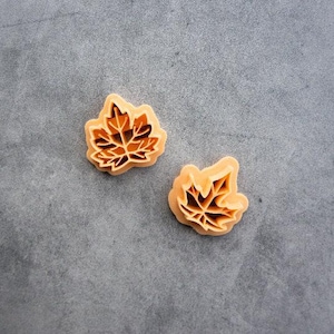 May include: Two light orange plastic cookie cutters shaped like maple leaves. Each cutter features a detailed interior vein pattern designed to imprint the leaf structure into dough, with a raised outer edge for cutting the leaf shape.