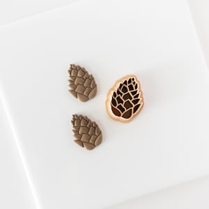 May include: Two small, light brown, textured pinecone-shaped cutouts made of clay or dough, positioned next to a single copper-colored pinecone-shaped cookie cutter with a detailed interior pattern for creating embossed impressions.