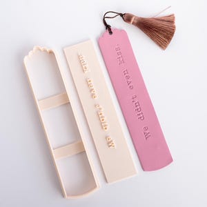 Op de afbeelding: Een roze bladwijzer met een bruin kwastje en de tekst We didn't even kiss. naast een beige plastic koekjesvorm en een bijpassende stempel met dezelfde tekst in een rechthoekige vorm met een afgeronde bovenrand.