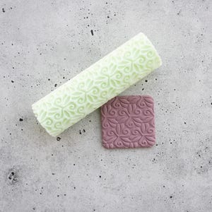 Pode incluir: Um rolo cilíndrico de cor verde-pálida com um padrão floral em relevo que se repete, colocado ao lado de uma peça quadrada de argila cor de malva escura que apresenta o mesmo design botânico intrincado e gravado na sua superfície.