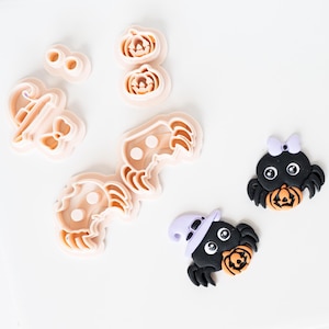 May include: A collection of light peach-colored plastic cookie cutters featuring shapes of pumpkins, a ghost, and spiders, alongside two finished spider decorations with black bodies, orange pumpkins, and purple accessories.