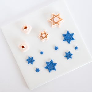 May include: A selection of star-shaped cookie cutters in different sizes and colours. The cutters are in blue, tan, and white. They are arranged on a white surface.