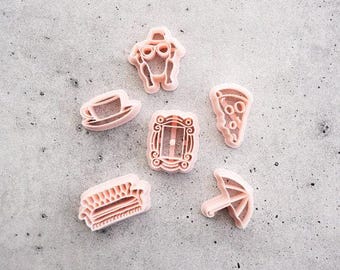 May include: A set of six light pink plastic cookie cutters featuring various shapes including a turkey with sunglasses, a coffee cup, a rectangular picture frame, a slice of pizza, a sofa, and an umbrella, all arranged on a flat surface.