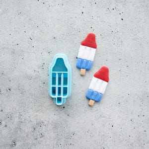 May include: A light blue plastic popsicle-shaped cookie cutter positioned next to two small, three-layered red, white, and blue popsicle-shaped decorations, each featuring a small wooden stick at the base.