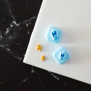 May include: Two blue plastic cookie cutters shaped like lightning bolts on a white surface. Two small gold cookie cutters are also on the surface.