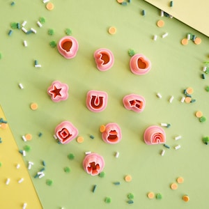 Op de afbeelding: Een verzameling van tien kleine, roze, plastic koekjesvormpjes in diverse vormen zoals een ster, hoefijzer, regenboog en andere geometrische figuren, verspreid gerangschikt naast kleine, kleurrijke, cilindervormige strooisels.