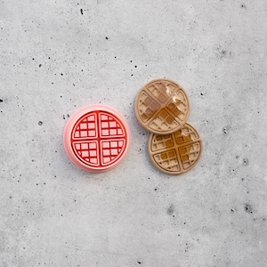 May include: A round pink waffle-shaped mold sits next to two small, circular, tan-colored waffle replicas. The replicas feature a grid pattern with a glossy, amber-colored syrup-like finish applied to the center of each individual waffle piece.