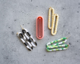 May include: Three pairs of elongated oval-shaped earrings and one pink plastic oval cutter. One pair is black and white with gold flecks, one is yellow with a white pattern, and one is green and white. Each earring has a gold-toned metal post.