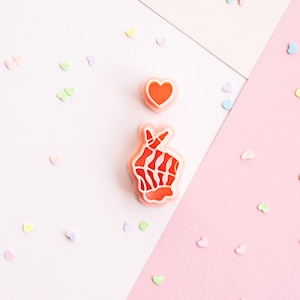May include: Two pink plastic cookie cutters on a pink and white background. The cookie cutters are shaped like a heart and a hand making the Korean heart sign. The background is covered in small pastel-colored hearts.