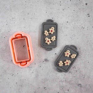 May include: A light pink rectangular cookie cutter with rounded handles and two grey rectangular trays, each featuring four small beige gingerbread-style figures with tiny red accents, arranged on a flat surface.