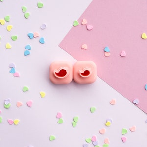 May include: Two pink heart-shaped cookie cutters with a red heart design on a light purple background with scattered pastel hearts.