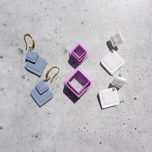 May include: Three pairs of geometric earrings with a layered design. One pair is light blue, one pair is white, and one pair is pink.