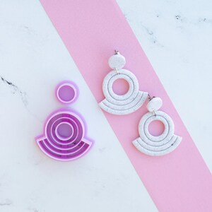 May include: A pair of white, speckled, circular earrings with a silver post and a silver ring in the center. The earrings are on a pink surface. Next to the earrings is a purple plastic mold for making the earrings.