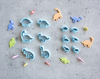 May include: A collection of light blue dinosaur-shaped cookie cutters alongside various small dinosaur-shaped stud earrings in pastel colors including pink, yellow, green, orange, and blue, arranged in a scattered pattern on a gray surface.