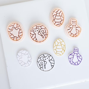 Puede incluir: Una colección de tres cortadores de galletas de plástico color melocotón claro con diseños de fantasma, gato y caramelo de maíz, junto a tres contornos correspondientes en blanco, negro y morado de las mismas formas sobre una superficie plana.