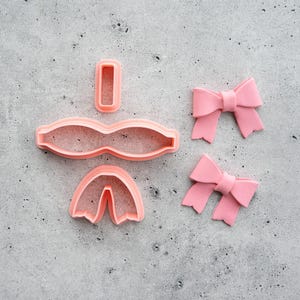 May include: A set of three light pink plastic cookie cutters designed to create a bow shape, including a rectangular centre piece, a wide curved top piece, and a bottom tail piece, alongside two finished light pink fondant bows.
