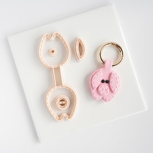 May include: A pink felt pig keychain with a gold-colored ring next to a pig-shaped cookie cutter and an ear-shaped cutter. The keychain has black button eyes and stitched details. The cookie cutters are light tan.