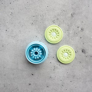 May include: A set of three blue and green cookie cutters shaped like flowers. The cutters are on a gray concrete background.