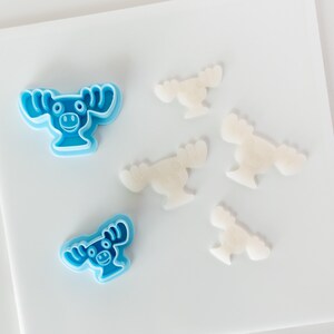 May include: Two light blue plastic moose-shaped cookie cutters and four white moose-shaped cutouts made of dough. The cookie cutters feature a detailed outline of a moose head with antlers, eyes, and a snout, while the dough cutouts are plain.