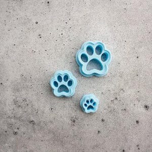 Pode incluir: Três cortadores de biscoitos azul-claros em forma de pegada de pata de tamanhos variados dispostos em grupo. Cada cortador apresenta uma almofada central e quatro dedos, com uma borda elevada para cortar massa ou outros materiais.