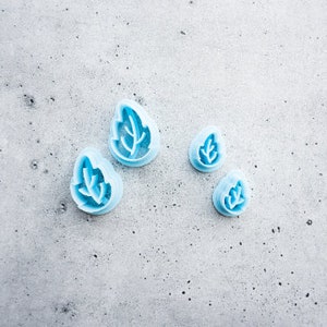 Może przedstawiać: Cztery niebieskie plastikowe foremki do ciasteczek w kształcie liści o różnych rozmiarach na szarej, teksturowanej powierzchni.