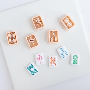 May include: A collection of five light peach-colored plastic mahjong tile cookie cutters and four white mahjong tiles with colorful engraved symbols in red, green, and blue. The set includes various character, bamboo, and circle suit designs.