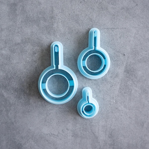 May include: Three light blue plastic cookie cutters in the shape of frying pans, arranged in a descending size order from largest to smallest. Each cutter features a circular base with a small rectangular handle extension at the top edge.