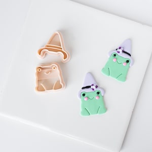May include: Two light peach plastic cookie cutters shaped like a frog and a pointed hat, alongside two finished light green frog figures wearing light purple pointed hats with small dark purple flower accents and pink cheeks.