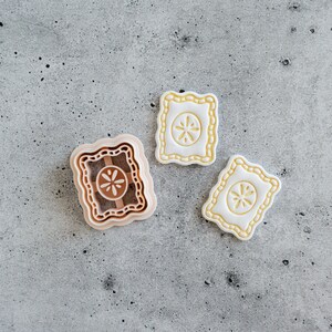 May include: A light peach-colored plastic cookie cutter with a scalloped rectangular frame and a central flower design, accompanied by two white rectangular cookies featuring the same scalloped edge and embossed flower pattern in the center.