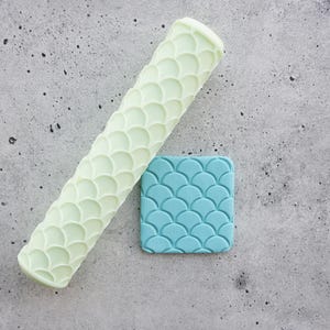 May include: A pale green cylindrical rolling pin featuring a repeating embossed fish scale pattern, positioned diagonally next to a square piece of blue material that displays the same embossed fish scale texture across its entire surface.