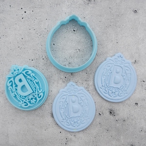 Könnte beinhalten: Drei blaue Plastik-Ausstecher, zwei mit einem dekorativen Design mit dem Buchstaben "B" und ein dritter einfacher runder Ausstecher.
