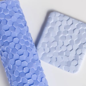 Puede incluir: Un rodillo cilíndrico de color azul bígaro claro y una baldosa cuadrada plana a juego, ambos con un patrón hexagonal de panal en relieve que se repite en toda su superficie, diseñado para crear impresiones texturizadas.