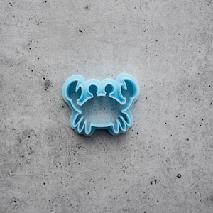 May include: A light blue plastic cookie cutter shaped like a crab. The cutter features a central body with two raised claws at the top and six legs extending from the sides, creating a clear outline for baking or crafting purposes.