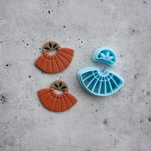Puede incluir: Dos pendientes de arcilla de color terracota con un dise&ntilde;o en forma de abanico y un peque&ntilde;o detalle de hoja. Junto a los pendientes hay un molde de pl&aacute;stico azul para hacer los pendientes.
