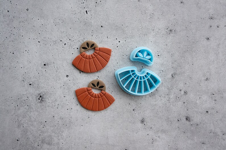 Puede incluir: Dos pendientes de arcilla de color terracota con un dise&ntilde;o en forma de abanico y un peque&ntilde;o detalle de hoja. Junto a los pendientes hay un molde de pl&aacute;stico azul para hacer los pendientes.