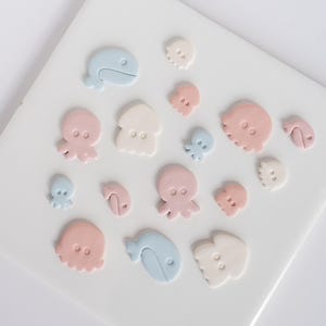 Pode incluir: Uma coleção de pequenos botões planos em forma de criaturas marinhas, incluindo baleias, polvos e lulas. Os botões apresentam uma paleta de cores suaves em azul pastel, rosa pálido e creme, cada um com dois pequenos orifícios.