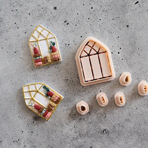 May include: Two decorated cookies shaped like a greenhouse with gold trim, featuring miniature plants and books. A matching cookie cutter and several smaller cutters are also present, all in a light pink color. The cookies are on a gray surface.